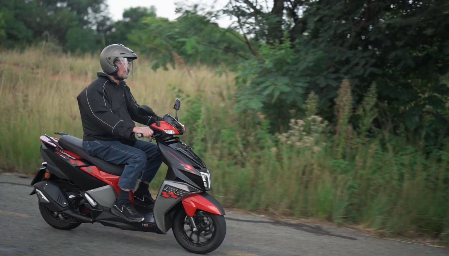 Still from Bike Test Drive: a rider on a red and black scooter on a road beside long grass